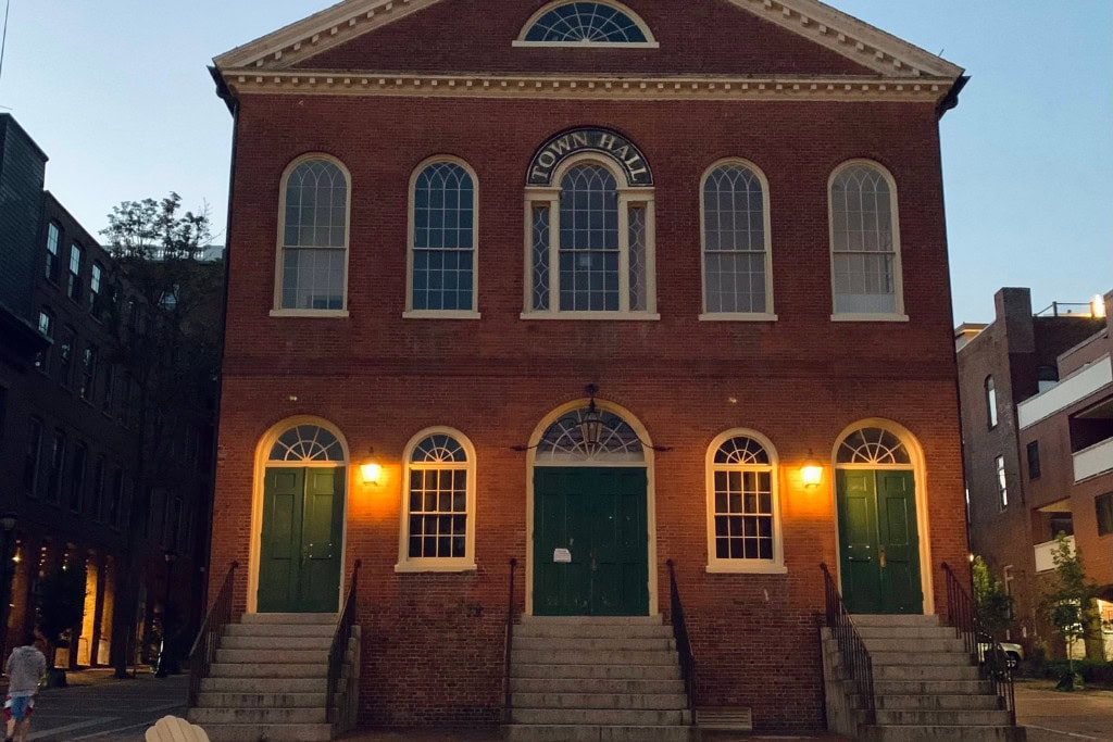 A historic brick town hall building illuminated by warm yellow lights at dusk, featuring symmetrical arched windows and three green double doors accessed by steps with black railings. The sky in the background is a soft gradient of light blue transitioning into night.