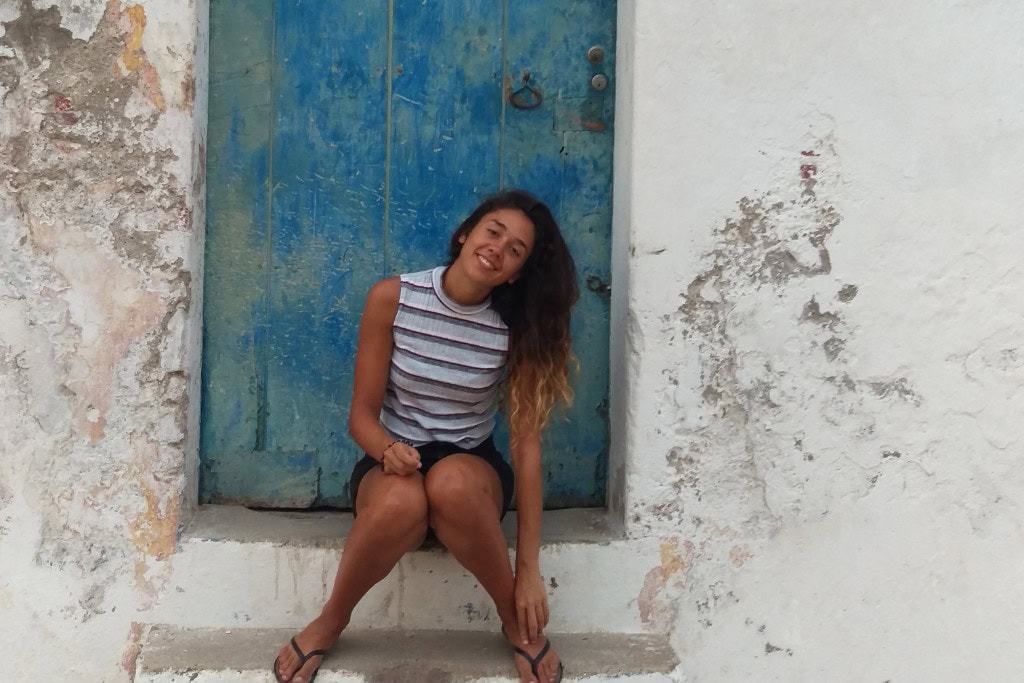 Huancabamba in Peru: Embark On A Spiritual Journey A woman sits on the steps of an old, weathered blue door set into a whitewashed wall, smiling warmly at the camera. The scene is taken in Canchaque, capturing a relaxed and rustic moment that reflects the charm of this small Peruvian town.