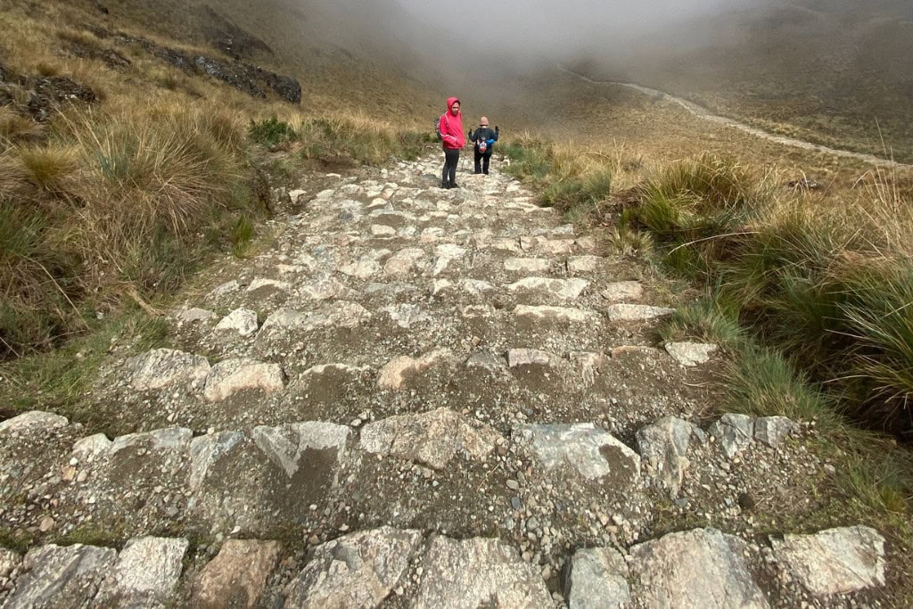 Huancabamba in Peru: Embark On A Spiritual Journey Two hikers walk along an ancient, stone-paved path winding through a highland landscape shrouded in mist, surrounded by rugged grasslands. This path, reminiscent of those built by the Inca civilization, evokes a deep connection to the culture's historical routes, highlighting the engineering and enduring legacy of the Inca Empire.