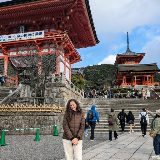 The image shows the author, Silvia, standing in front of the vibrant red gate of Kiyomizu-dera Temple in Kyoto, Japan, surrounded by other tourists and stone steps leading up to the main temple complex. The iconic pagoda is visible in the background against a cloudy sky, capturing the lively atmosphere and architectural beauty of this popular historical site. The scene reflects Kyoto’s cultural significance and appeal to international visitors.
