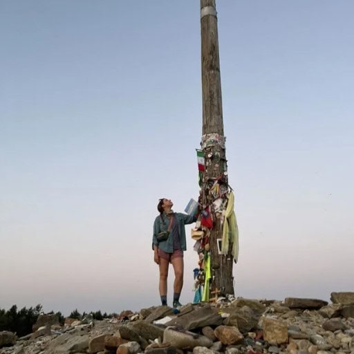 This image shows a person, Mikala Whitaker, standing next to a tall wooden pole adorned with various items such as scarves, ribbons, and flags. The person is looking up at the pole, which stands on a rocky ground, under a clear sky, creating a scene that suggests reflection or a moment of achievement.