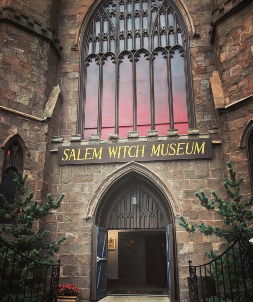 Personal Visit To Salem Massachussets Witch Village: Everything You Need to Know! The entrance of the Salem Witch Museum, showcasing its grand stone facade with tall, arched stained glass windows and an open wooden double door. The museum's name is prominently displayed on a sign above the entrance, set against a backdrop of rich, weathered stone, flanked by small evergreen trees.
