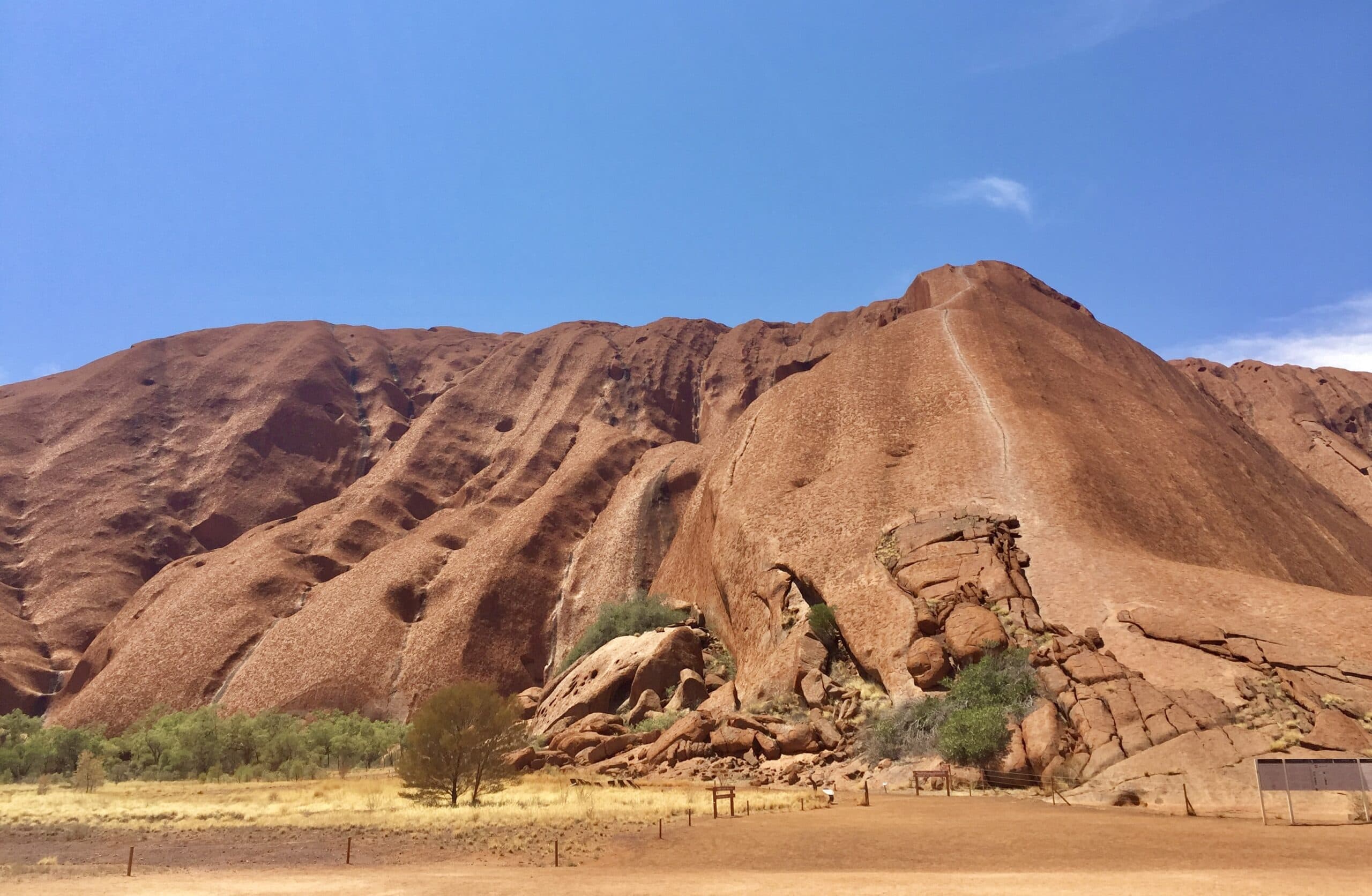 Uluru under a clear blue sky, showcasing its iconic red sandstone and spiritual significance. The rock’s surface features natural grooves and erosion patterns, surrounded by sparse vegetation and a flat, arid landscape, reflecting Uluru's cultural and spiritual value.