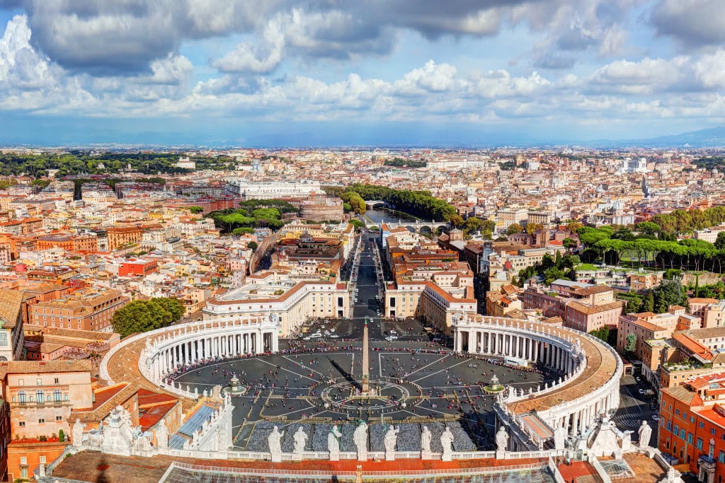 Spiritual Quest St. Peter Basilica (Vatican City) A panoramic view of St. Peter's Square in Vatican City, featuring its iconic colonnades, central obelisk, and statues, with the city of Rome sprawling into the distance under a partly cloudy sky.