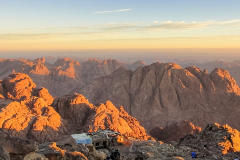 Golden sunlight illuminating rugged mountain peaks, with a small structure nestled among the rocks and a vast horizon stretching into the clear sky.