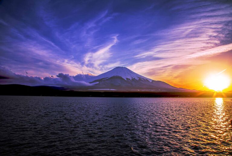 A vibrant sunset over Mount Fuji, with its snow-capped peak silhouetted against a dramatic sky of purple, orange, and yellow hues, reflected on the calm waters below.