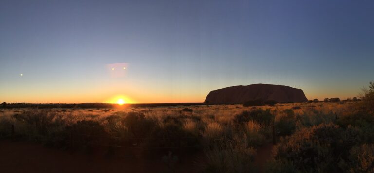 Panoramic view of Uluru at sunrise, with golden sunlight casting a warm glow over the desert landscape and grasslands. The iconic rock formation stands majestically under a clear sky transitioning from deep blue to light orange.