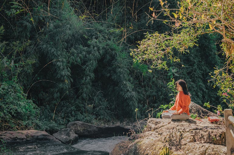 Image of a woman meditating in a seated position by a serene riverside, surrounded by lush greenery. She is bathed in warm sunlight, with the calm water reflecting the vibrant hues of the surrounding trees and plants, creating a peaceful and tranquil atmosphere. Image used for the article Spiritual Manifestation.