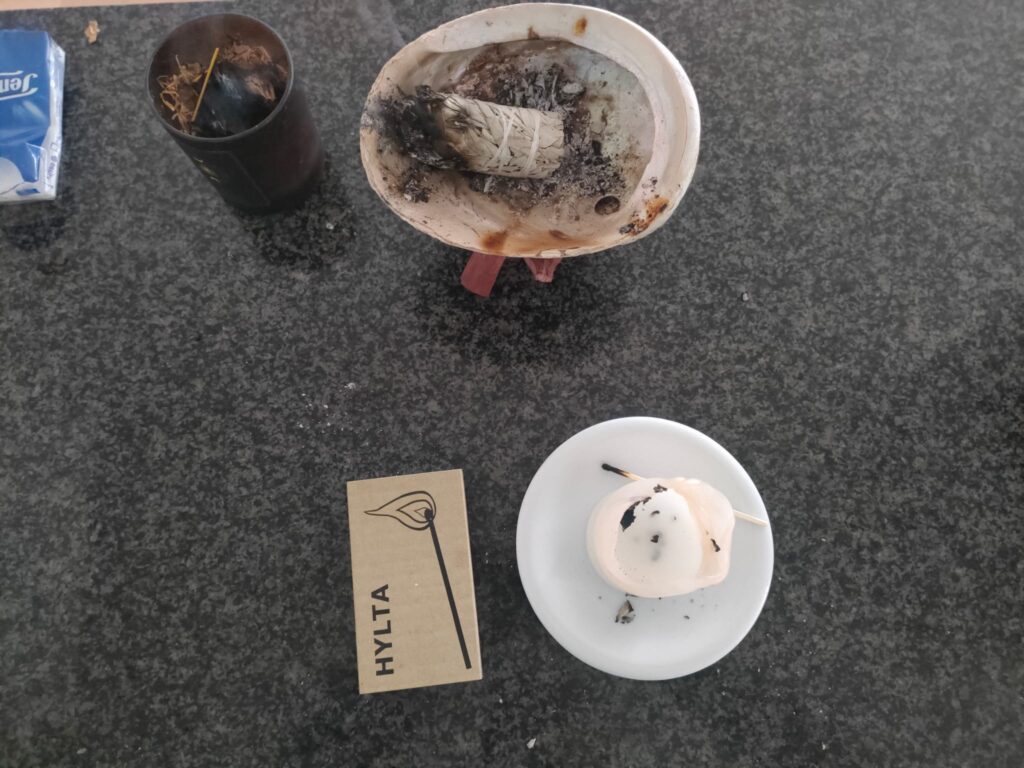 How to Energetically House Cleansing with Sage? A dark granite countertop displays various spiritual and ritualistic items. At the top, a white ceramic or shell bowl holds a partially burned sage bundle with ashes scattered around. To the left, a blackened cup contains herbs and incense sticks, emitting smoke. Below, a beige card with a minimalist drawing of a burning plant stem and the word "HYLTA" rests on the countertop. On the right, a melted candle with a burnt wick sits on a small white plate, with blackened residue around it. The scene suggests a smudging or cleansing ritual.