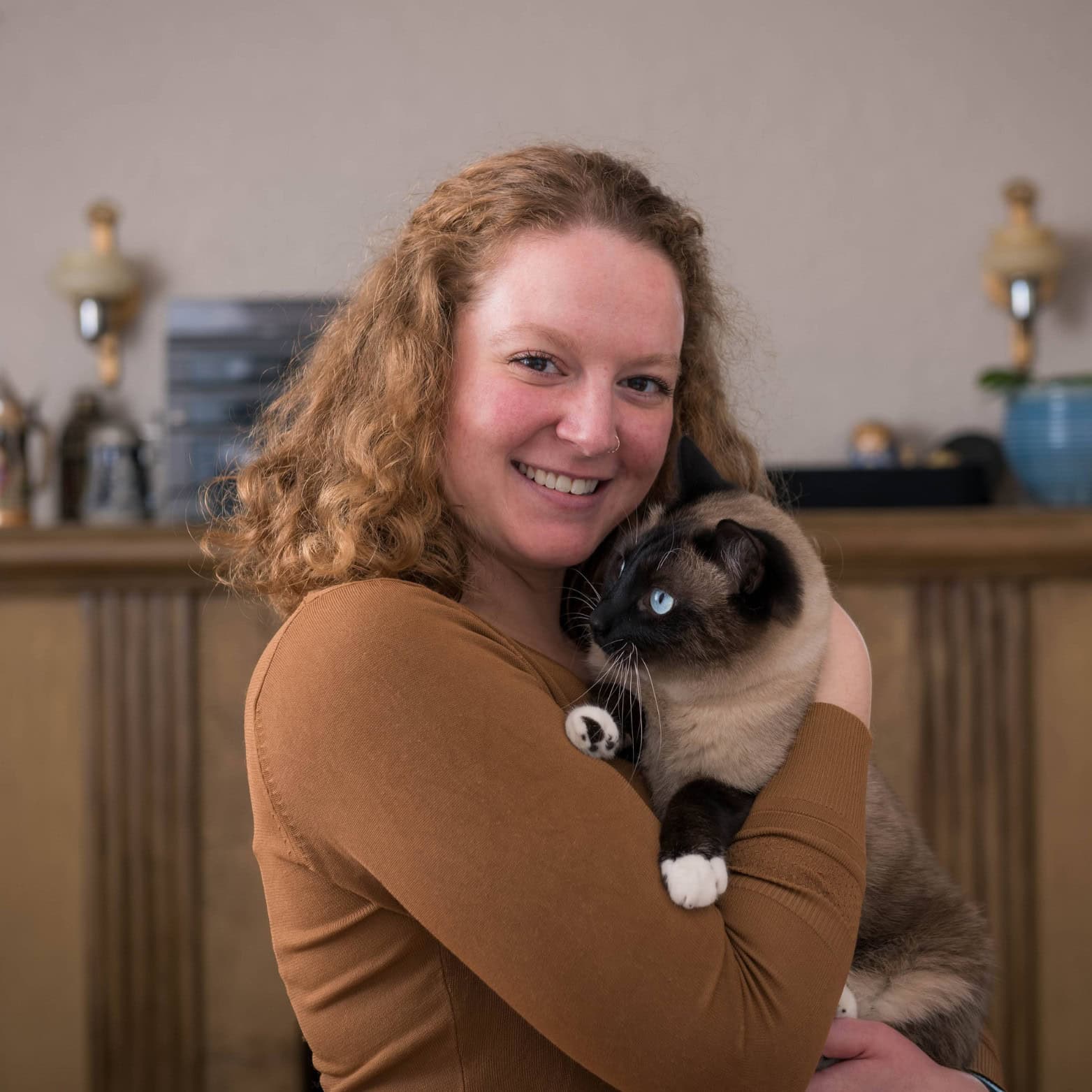 The Leshan Sprit Guide to the Leshan Giant Buddha A woman, Moly Clayton, with curly red hair wearing a rust-colored top smiles while holding a Siamese cat with bright blue eyes and white-tipped paws. The cozy indoor setting features a wooden mantel with decorative objects in the background.