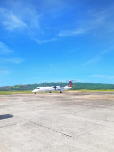 A small Philippine Airlines propeller plane parked on the runway at Coron Palawan airport, surrounded by a wide concrete tarmac under a bright blue sky. In the background, green hills and tropical vegetation rise gently, highlighting the remote and natural setting of Coron Palawan. This image captures the typical arrival experience for travelers starting their 3-day trip to Coron Palawan, emphasizing the island’s accessibility and beautiful landscape upon landing.