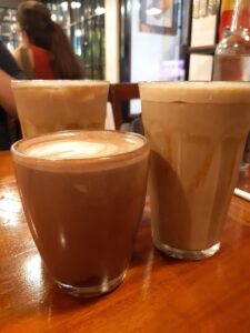 Close-up of three coffee drinks on a wooden table in a café in Coron Palawan, featuring one cup of hot latte with foam art and two tall glasses of iced coffee, creating a cozy and inviting atmosphere during a 3-day trip to Coron Palawan.