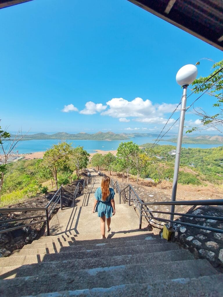 Woman walking down the stairs of Mount Tapyas in Coron Palawan on a sunny day, with a panoramic view of green hills, the ocean, and blue sky, capturing the beautiful landscape during a 3-day trip to Coron Palawan.