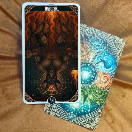 Image of The Devil Tarot Card in reversed position. The card shows an inverted dark silhouette with a white glowing circle at its feet, chains hanging upward, and red flames extending downward. The background features a cosmic space filled with stars and planetary symbols. The card rests on a brown velvet surface, with part of a colorful card back underneath showing swirling patterns of blue, red, green, and yellow, with the word 'Sabiduri' in the center.