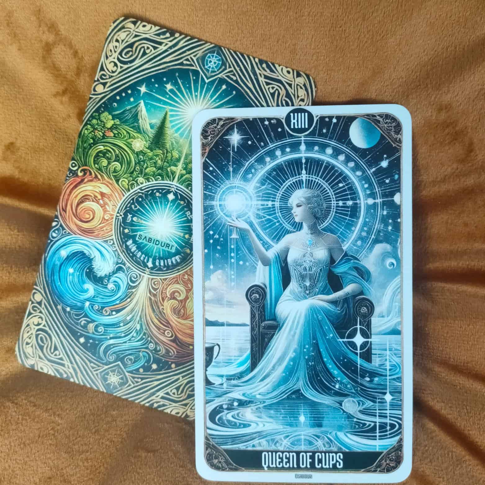 The Queen of Cups tarot card in upright position, placed on a brown cloth with the Sabiduri deck’s ornate back design visible. The illustration shows a regal woman in flowing blue robes seated on a throne by the ocean, holding a glowing sphere, symbolizing compassion, intuition, and emotional balance.