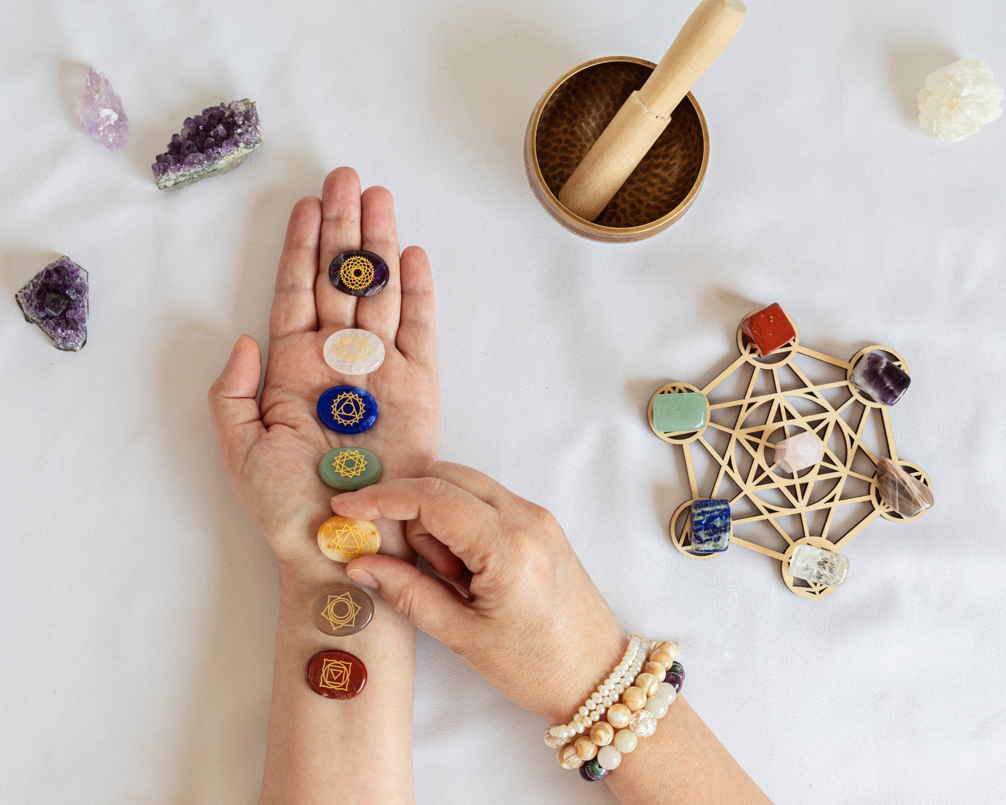 A hand displaying seven chakra stones arranged along the arm beside a wooden crystal grid, amethyst clusters, and a singing bowl on a white surface.