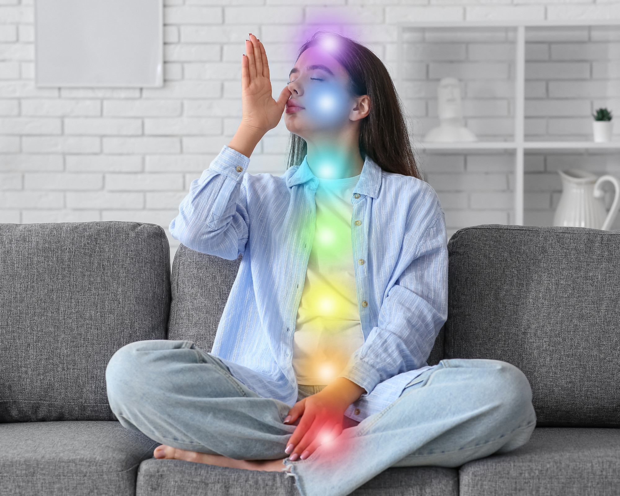 A woman sitting cross-legged on a sofa practicing alternate-nostril breathing while glowing chakra colors align from the root to the crown along her body.