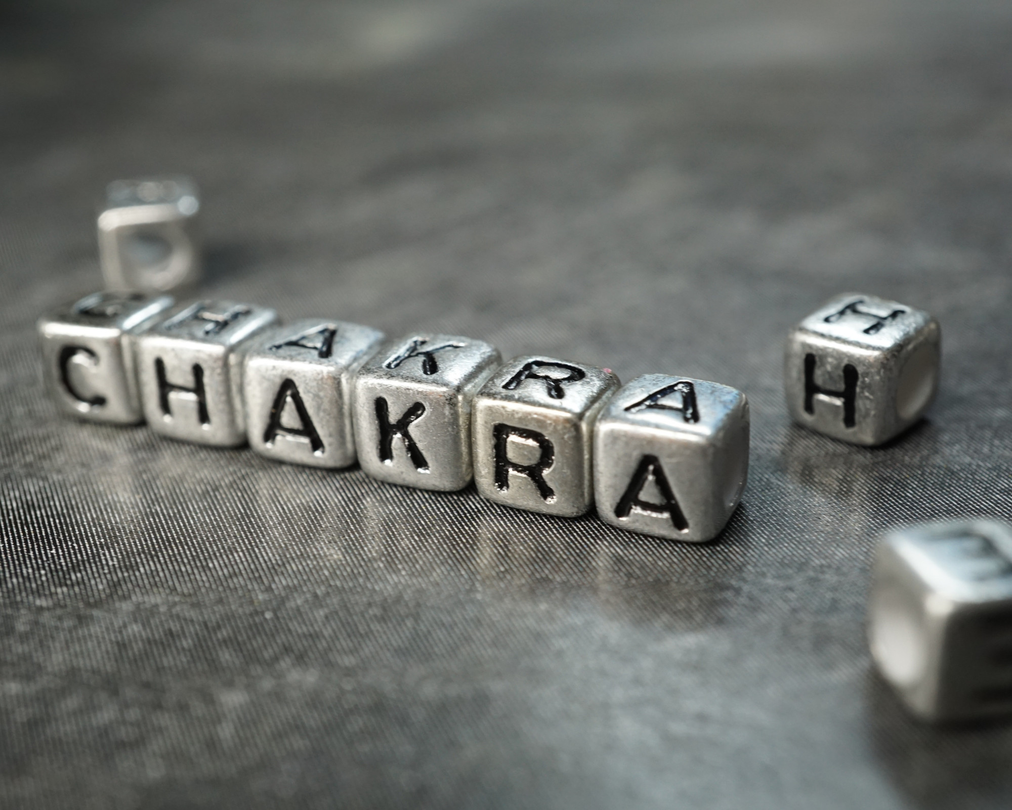 Close-up of metallic letter cubes spelling the word ‘Chakra’ on a textured gray surface."
