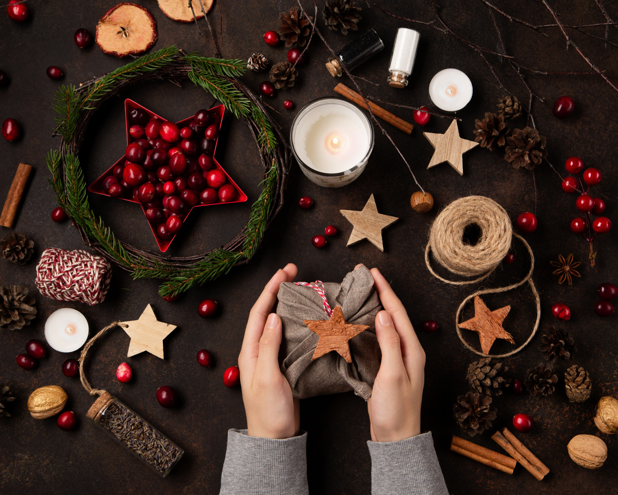 Hands preparing handmade Yule gifts surrounded by pinecones, cranberries, candles, twine, and natural decorations, showing creative ideas for how to celebrate Yule with crafting and intention.