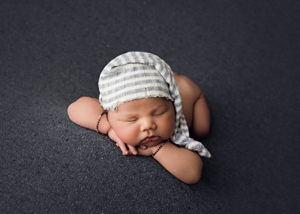 Sleeping newborn baby boy wearing a blue and white sleepy hat, posed with his hands beneath his face
