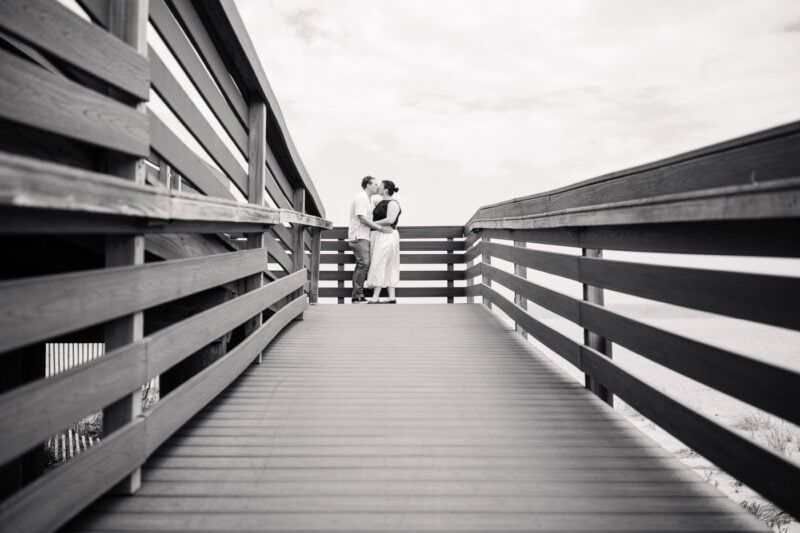 The Perfect Black & White Photo Editing Style Couple embracing on wooden boardwalk under cloudy sky.
