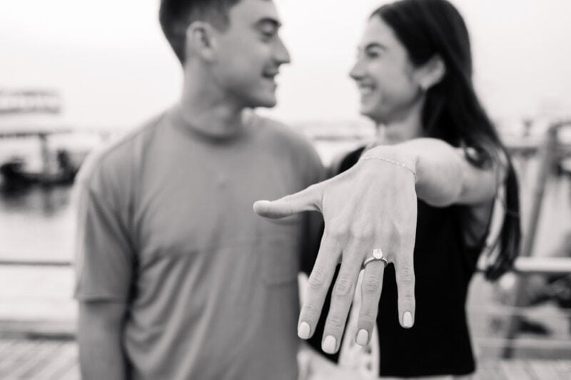 The Perfect Black & White Photo Editing Style Engagement ring close-up with smiling couple in background.