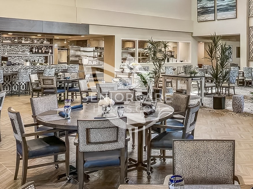Main dining room at Clearwater at Pinnacle Peak with round tables, patterned upholstered chairs, potted palms, and an open display kitchen.