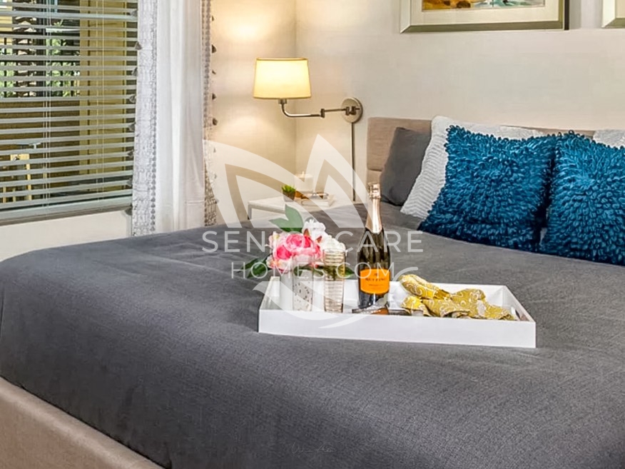 Apartment bedroom at Clearwater at Pinnacle Peak with a gray bedspread, teal accent pillows, a wall-mounted reading lamp, and a breakfast tray with flowers.