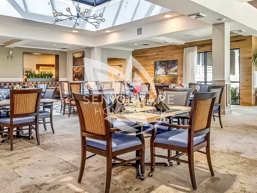 Dining room at Clearwater at North Tustin with a skylight, cane-back chairs, a wood accent wall, and tables set with fresh flowers.