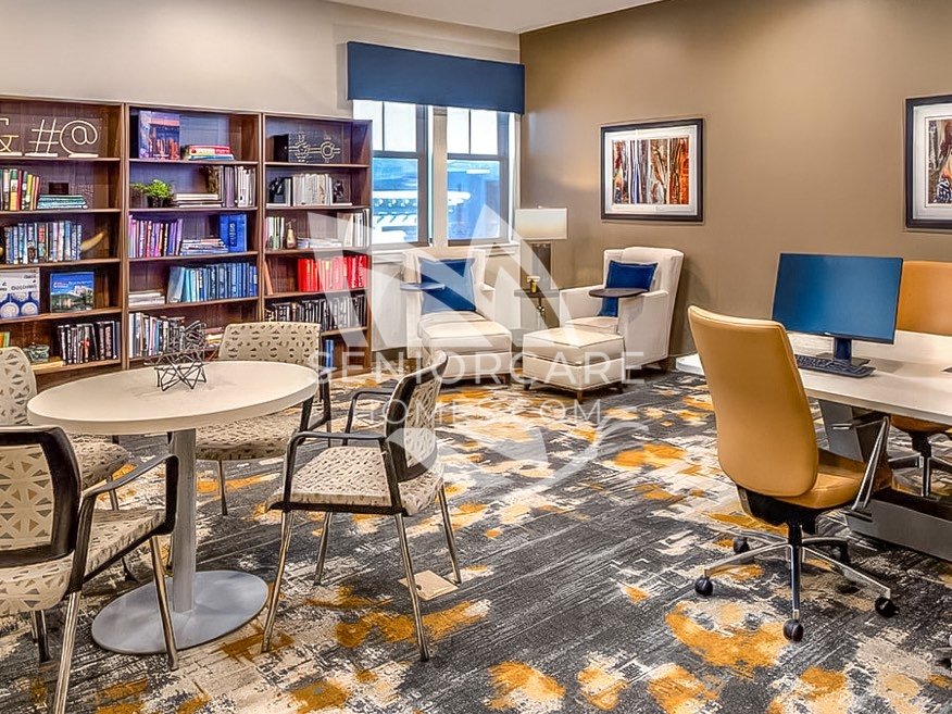 Library and computer room at Clearwater at Rancharrah with full bookshelves, a round table, cream reading chairs, and a desktop computer station.