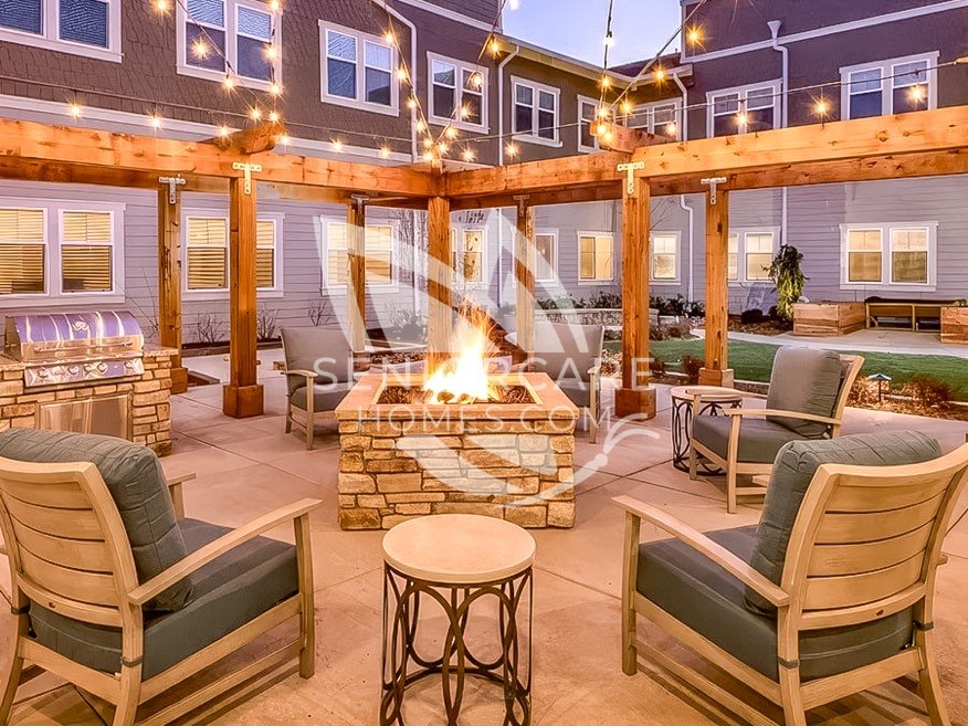 Courtyard at Clearwater at Rancharrah at dusk, with a stone fire pit, cushioned lounge chairs, a wooden pergola with string lights, and a built-in grill.