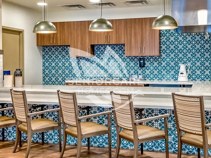 Bistro counter at Clearwater at Rancharrah with a teal patterned tile backsplash, wood cabinets, upholstered counter chairs, and brass pendant lights.