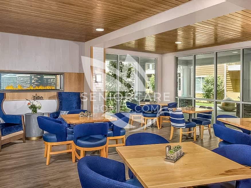 Casual dining area at Clearwater at Riverpark with blue tub chairs, wood tables, a linear fireplace, and floor-to-ceiling windows facing the courtyard.