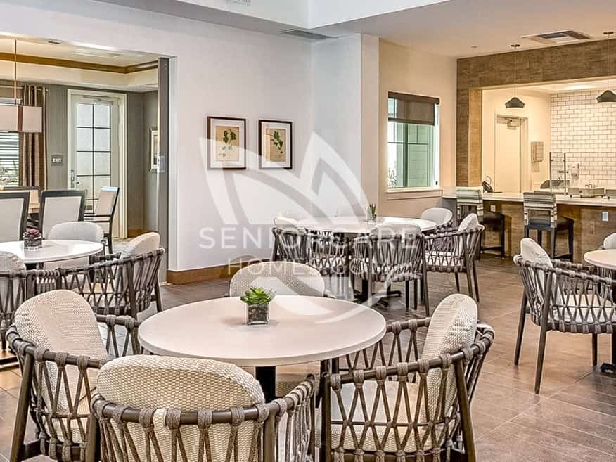 Dining area at Clearwater at Sonoma Hills with round white tables, woven rope chairs, botanical prints, and a serving counter with white subway tile.