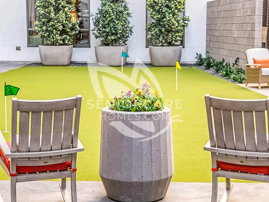 Courtyard putting green at Clearwater at The Heights with colorful flags, a planter of pansies, and two wooden rocking chairs with red cushions.