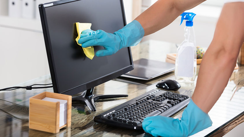 Person cleaning a computer monitor with a yellow cloth, wearing blue gloves.