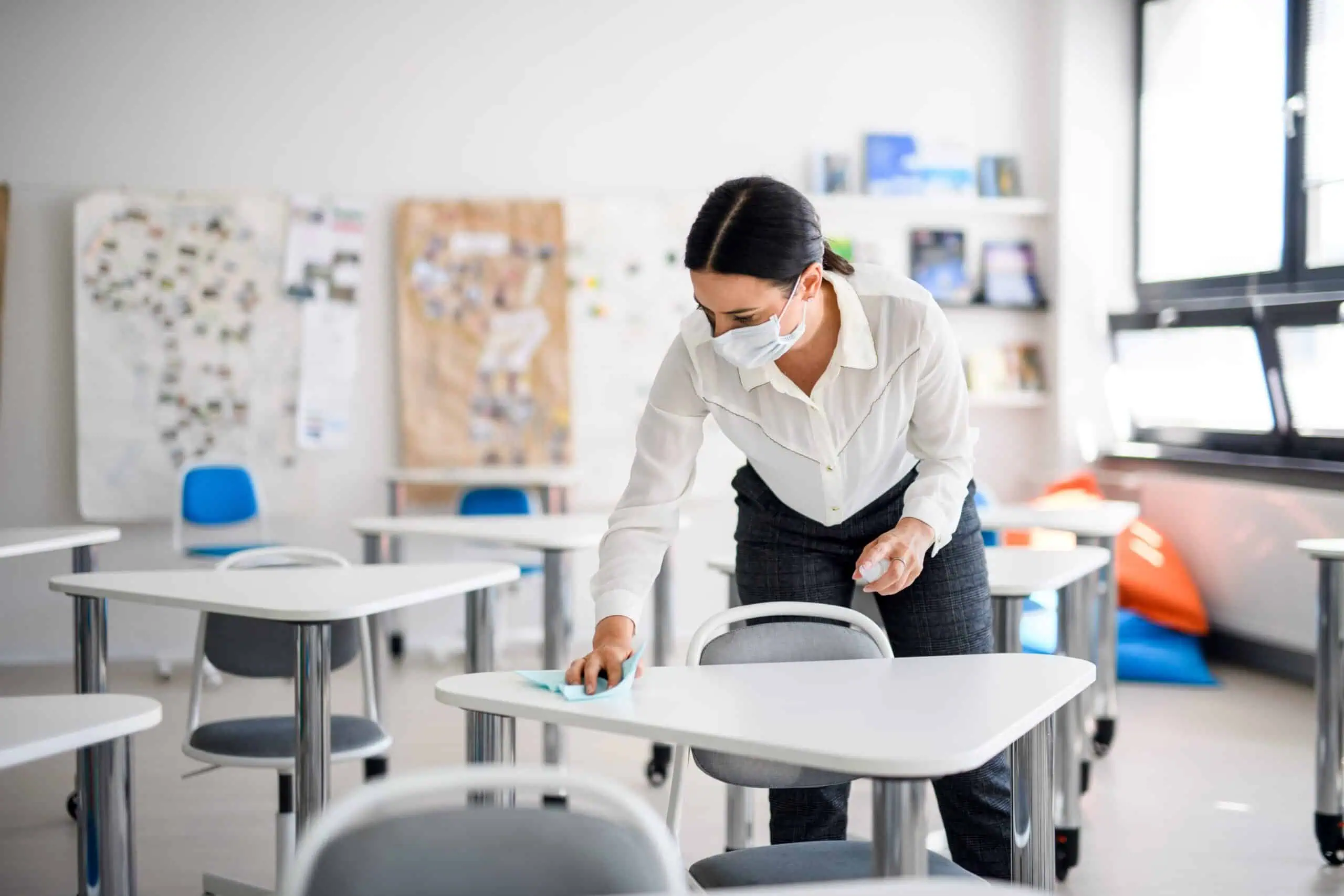 Woman wearing face mask cleaning classroom desks with cloth.