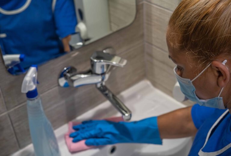A cleaning professional wearing gloves and mask disinfects a sink in a medical setting.
