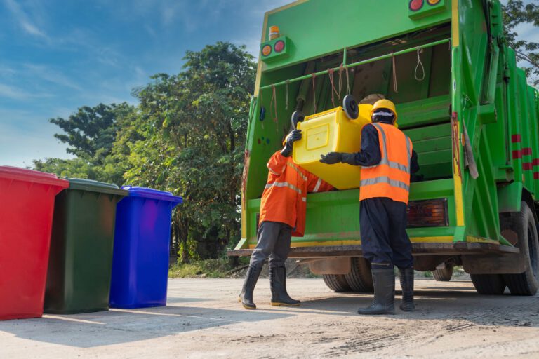 Two waste collection workers in safety vests and helmets empty a yellow bin into a green garbage truck