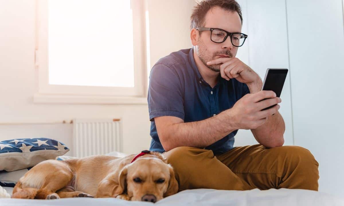man with brown hair and glasses, wearing a dark blue polo shirt and khaki pants sitting on a bed looking at his cellphone with a golden brown dog laying beside him
