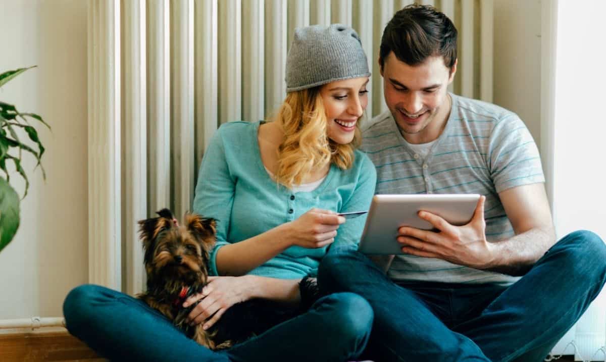 A young couple is sitting cross-legged on the floor next to each other, both looking at a tablet in his hand. She is holding out a credit card, with a small brown dog in her lap. She is wearing a blue shirt, a grey beanie hat over her blonde hair and blue jeans. He is wearing a grey striped shirt and blue jeans.