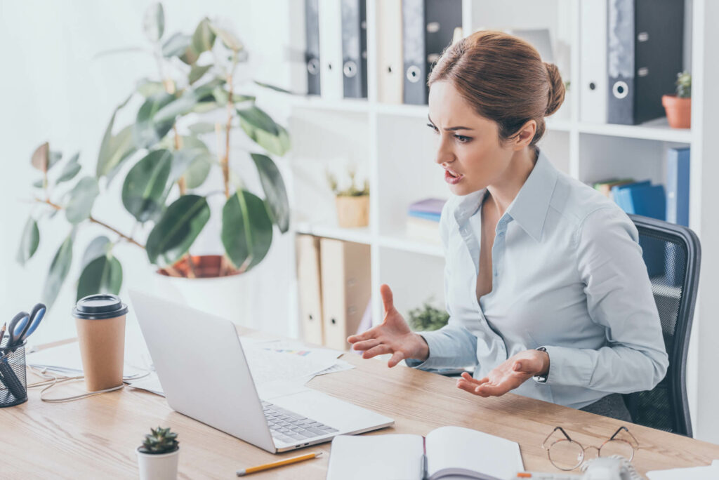 A teacherpreneur sits at a desk with an open laptop, appearing frustrated and gesturing with her hands. A coffee cup, papers, and a notebook are scattered across the desk. In the background, bookshelves and plants create a cozy workspace.