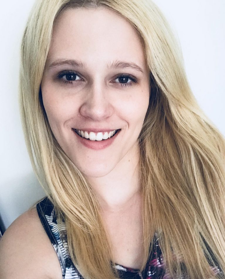 A person with long blonde hair is smiling at the camera, wearing a patterned sleeveless top, in front of a plain light background.