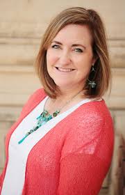 Lisa Vangemert, a woman with shoulder-length light brown hair, wearing a coral cardigan, white top, and turquoise necklace, smiles at the camera with a neutral background.