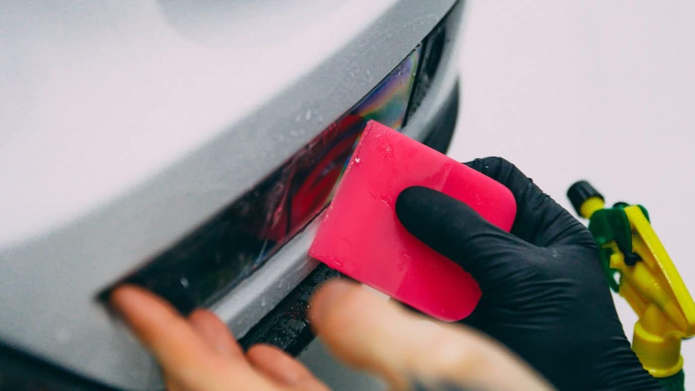 Close-up of hands applying red protective film to car's rear bumper.