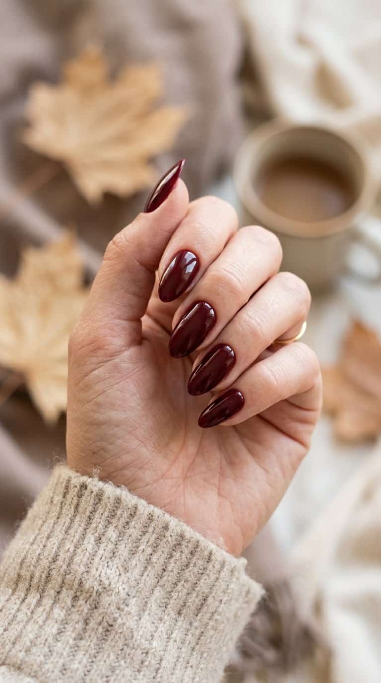 A hand with glossy deep burgundy almond-shaped nails rests against a warm autumn-toned background.