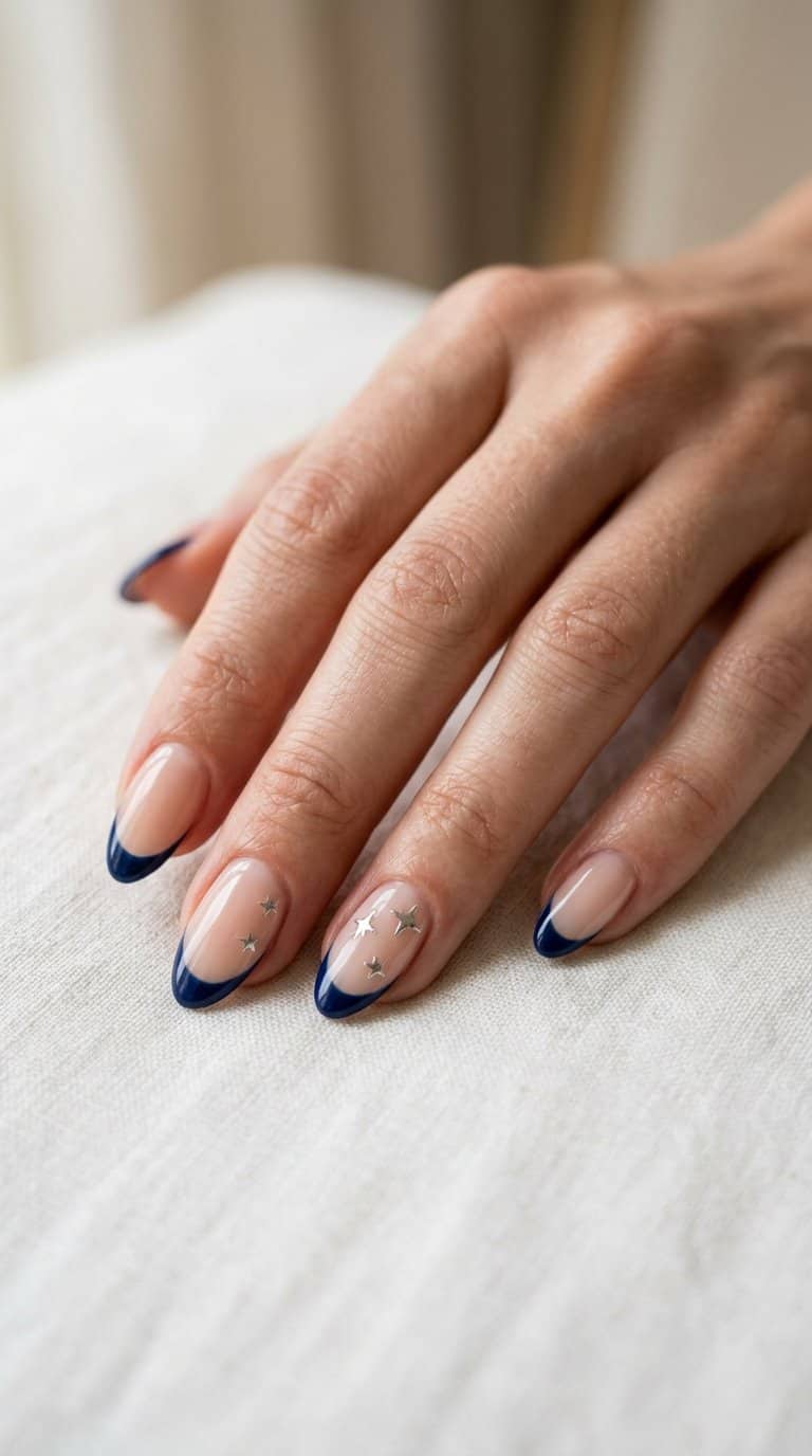 A hand with glossy nude nails featuring navy French tips and small silver star accents.