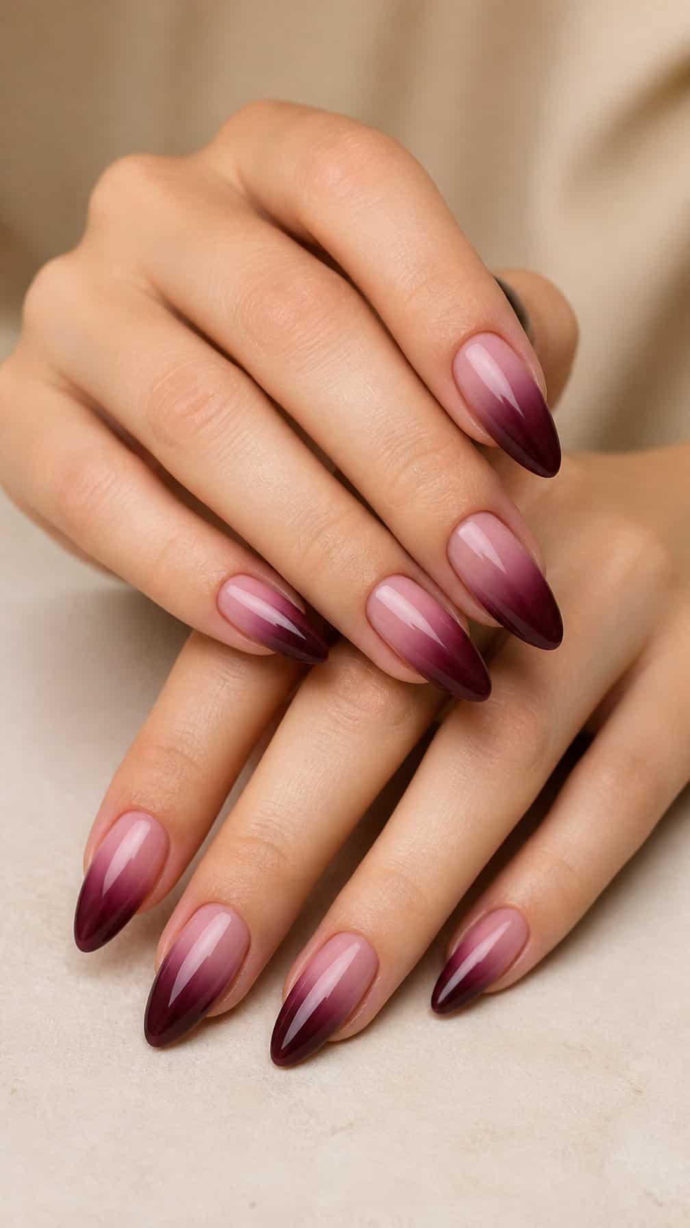 A close-up of a hand with glossy almond-shaped nails transitioning from deep burgundy to soft blush pink.