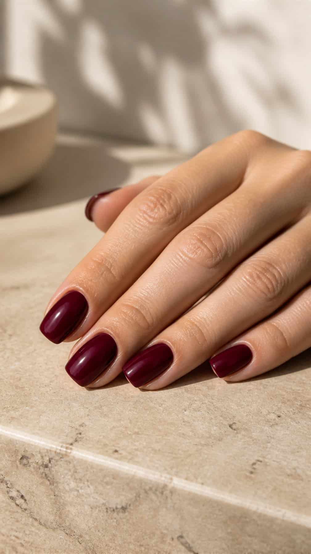 A hand displays glossy burgundy square-shaped nails on a neutral surface.