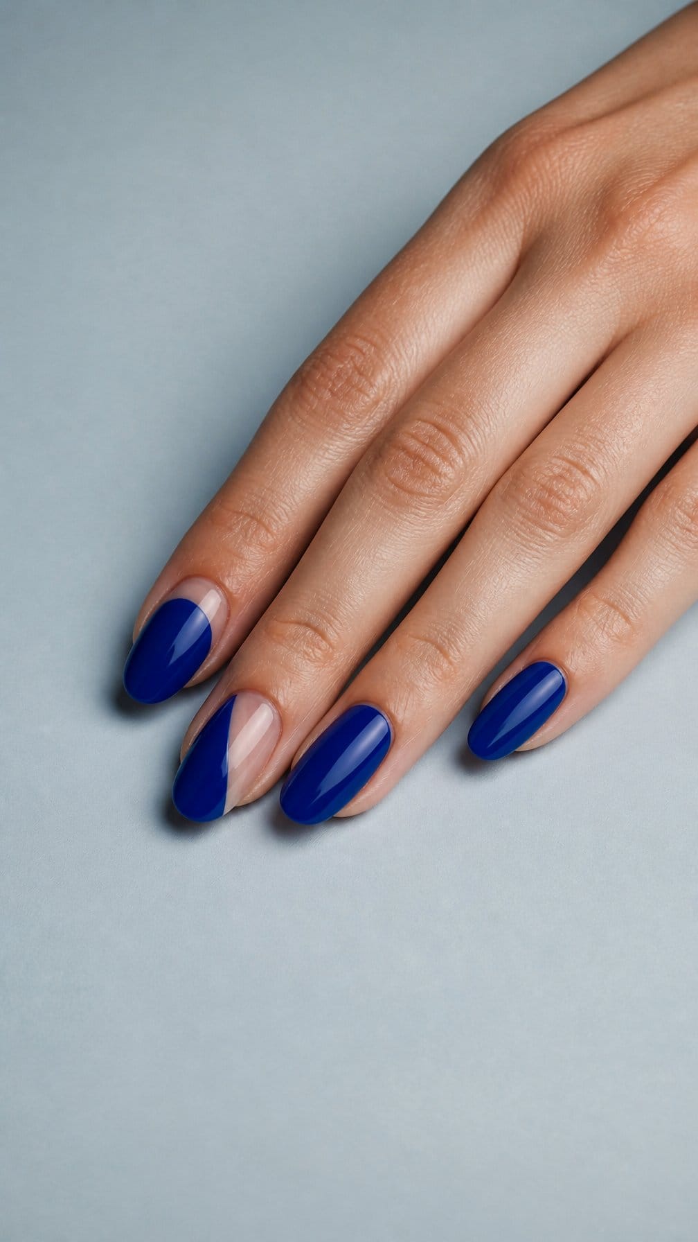 A hand with a glossy cobalt blue manicure featuring minimalist bare-nail accents rests on a pale surface.